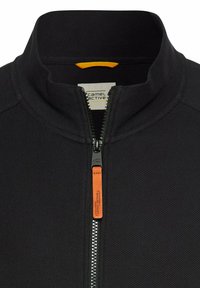 Zwarte zip-up jas van een textuurstof, met hoge kraag, oranje ritslabel en een logotag nabij de kraag. Ritsdetails zijn opvallend.