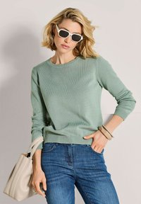 Blonde Frau mit weißen Sonnenbrillen, grünem Pullover, blauen Jeans, beigem Armband und einer beigen Handtasche, die vor einem hellen Hintergrund steht.