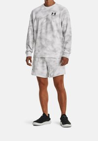 Hombre vestido con sudadera de manga larga en camuflaje blanco y gris y pantalones cortos a juego, con zapatos deportivos negros, de pie frente a un fondo blanco.