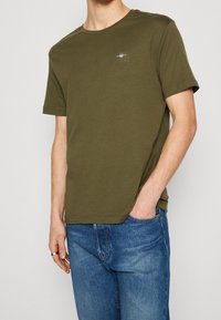 T-shirt in cotone verde oliva con scollatura rotonda e maniche corte. Presenta un piccolo logo ricamato sul petto. Indossato con jeans in denim blu.