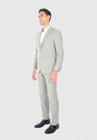 Completo grigio chiaro con vestibilità su misura, giacca a un bottone, rever a punte e tasche laterali, abbinato a scarpe nere. Camicia bianca sotto.