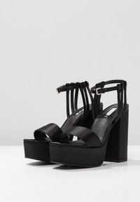 Černé platformové sandály se saténovým svrškem, otevřenou špičkou a nastavitelnými nártními pásky. Blokové podpatky poskytují stabilitu. Hladká textura a jednoduchý design.