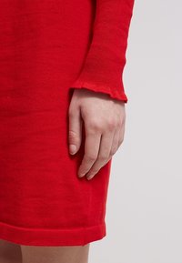 Abito rosso lavorato a maglia con una silhouette aderente. Le maniche lunghe presentano un orlo ondulato. Mano mostrata appoggiata sul tessuto testurizzato dell'abito.