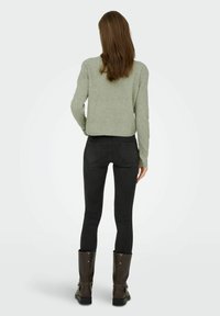 Pelēki zaļš piegriezuma džemperis ar garām piedurknēm, apvienots ar melnajām pieguļošajām džinsām un brūnajām līdz ceļiem zābakiem, skatīts no aizmugures.