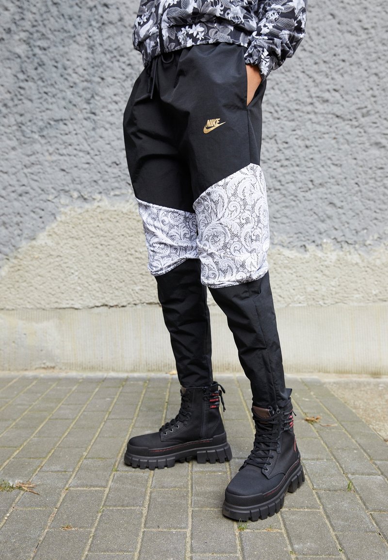 Černé kalhoty s bílým paisley panelem a elastickým pasem. Černé robustní šněrovací boty s texturovanou podrážkou. Zlaté logo Nike.