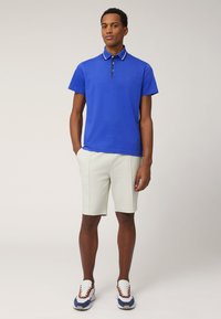 Polo shirt blu royal con strisce bianche sul colletto, abbinato a pantaloni corti beige chiaro. Il modello indossa sneakers bianche e blu.