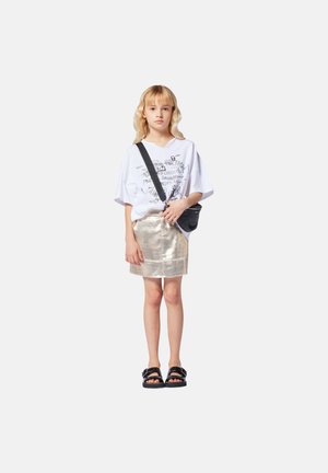 T-shirt ample blanc avec impression graphique, mini jupe dorée métallique, sac bandoulière noir, et sandales noires à larges brides.