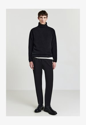 Schwarzer gestrickter Rollkragenpullover, strukturierter Stoff, kombiniert mit engen schwarzen Hosen und schwarzen Ankle Boots. Sauberes und minimalistisches Design.