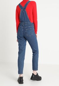 Salopette di denim blu scuro con bretelle regolabili incrociate sulla schiena, gambe affusolate e due tasche posteriori. Indossata con una maglia rossa a maniche lunghe e scarpe nere.