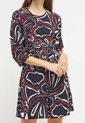 Frau trägt ein langärmliges Kleid mit roten, schwarzen und weißen abstrakten Blumen- und geometrischen Mustern.