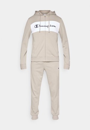 Lysebeige træningssæt i blødt stof med en hvid panel med navyblåt "Champion"-logo på hættetrøjen. Inkluderer elastiske manchetter og linning.