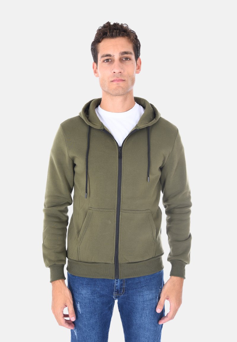 Felpa con zip verde oliva realizzata in tessuto morbido, con lacci neri, tasche frontali e polsini a coste, indossata sopra una maglietta bianca.