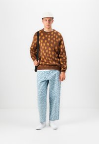 Jonge man in een bruine gestippelde trui, lichtblauwe polkadotbroek, witte sneakers, witte pet en zwarte schoudertas, staande tegen een witte achtergrond.