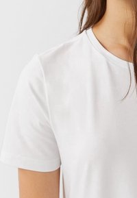 Gros plan sur une personne portant un t-shirt blanc uni à manches courtes, avec des cheveux bruns raides sur l'épaule, contre un fond blanc.