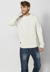 Fehér bordás kötött pulóver texturált mintával, kerek nyakkivágással és hosszú ujjal, sötétkék farmerral párosítva.