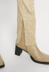 Béžové kožené kotníkové boty se čtvercovou špičkou a masivním černým podpatkem, doplněné o texturované béžové kalhoty s bočními rozparky.