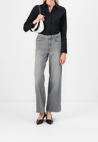 Camicia nera con bottoni, jeans grigi a vita alta e a gamba larga, décolletés neri a punta, e una piccola borsa bianca con dettagli neri.