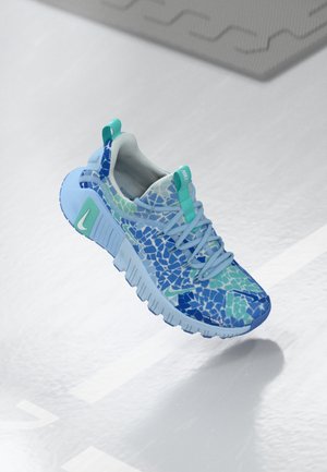 Zapatilla azul claro con patrón de mosaico en azul y verde azulado, lengüetas de tirón verde azulado y suela gruesa segmentada, flotando sobre una superficie blanca brillante.