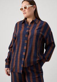 Raiščiuotas mygtukų marškiniai jūros mėlynos ir rūdžių spalvos, su laisvu pritaikymu, ilgomis rankovėmis ir minkšta tekstūra. Derinami su kampuotais baltais saulės akinių.