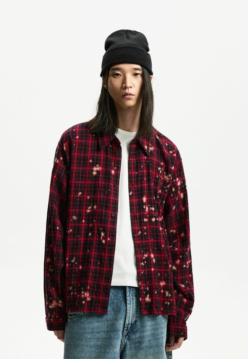 Chemise en flanelle à carreaux rouges et noirs avec des taches de javel, dotée d'un devant boutonné et de manches longues, portée par-dessus un T-shirt blanc et un jean bleu.