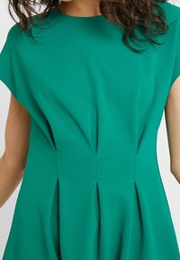 Grön klänning i mjukt tyg med en slät textur, kapsleeves och vertikala veck längs bodicen, vilket skapar en strukturerad design.