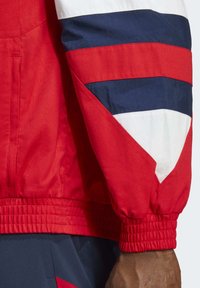 Chaqueta ligera roja con acentos geométricos en blanco y azul marino, puños elásticos y dobladillo ajustado para un ajuste ceñido. Tejido texturizado.