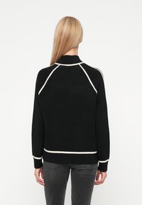 Melnā adīšana džemperis ar baltu apdari, ar augstu apkakli un raglanu piedurknēm, redzams no aizmugures. Saskaņots ar tumši pelēkām biksēm.