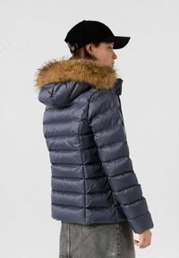 Chaqueta de plumas azul marina con capucha forrada de piel sintética, diseño acolchado horizontal y cierre de cremallera. La tela parece suave y brillante.