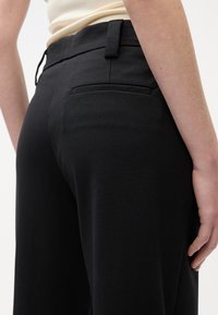 Gros plan d'une personne portant un pantalon noir sur mesure avec une poche passepoilée à l'arrière et des passants pour ceinture, debout devant un fond clair uni.