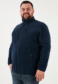 Tamsiai mėlynas fliso džemperis su aukštu apykakle, pilnu priekiu užtraukiamu užtrauktuku ir dviem šoniniais kišenėmis. Yra užtrauktuku užsegama krūtinės kišenė ir minkšta tekstūra.