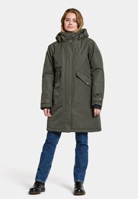 Olivgrüner langer Parka mit Kapuze, Reißverschluss und Druckknöpfen, zwei Fronttaschen und verstellbaren Bündchen, getragen über blauen Jeans und Stiefeln.