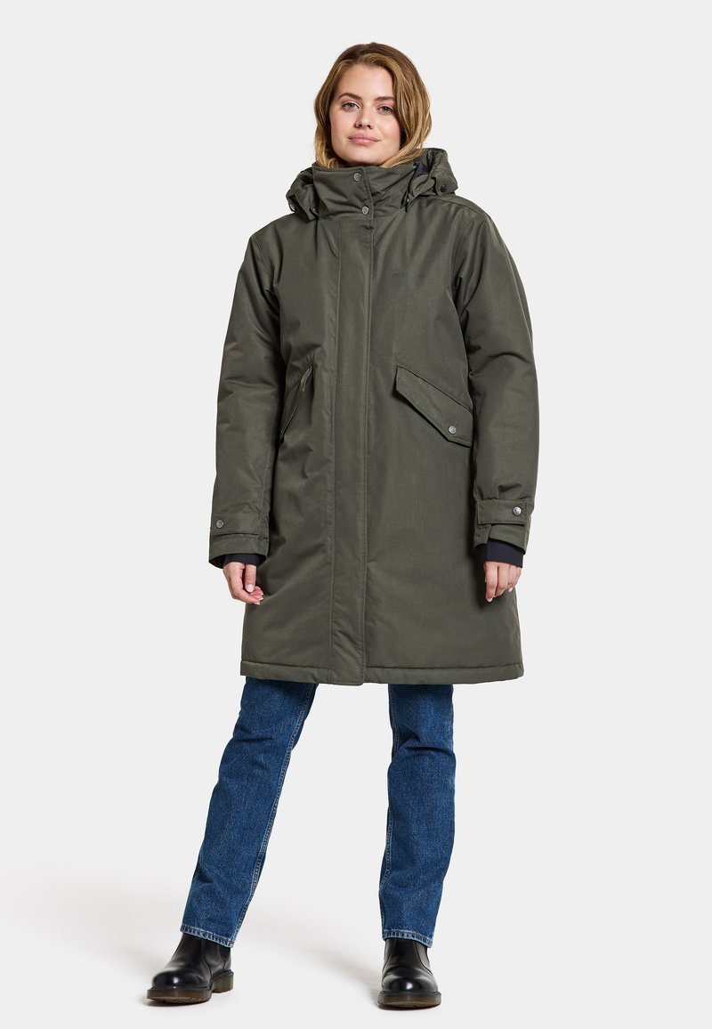 Olivgrüner langer Parka mit Kapuze, Reißverschluss und Druckknöpfen, zwei Fronttaschen und verstellbaren Bündchen, getragen über blauen Jeans und Stiefeln.