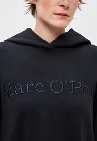Čierna ľahká mikina s kapucňou s jemnou textúrou. Na prednej strane je vyšité názov značky "Marc O'Polo" tmavomodrou farbou.