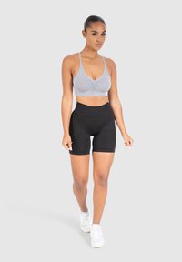 Grijze sportbeha met een gerimpelde voorkant, gedragen met hoge zwarte shorts. Model draagt witte sneakers en staat tegen een neutrale achtergrond.