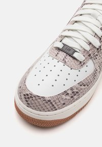 White and snakeskin-patterned sneaker with perforated toe, textured accents, and gray rubber sole. Laces and logo in dark beige.