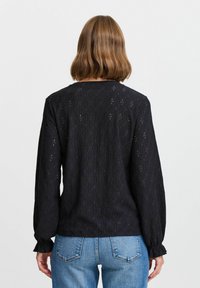 Top en dentelle noire avec un motif texturé, manches longues et poignets froncés, associé à un jean en denim bleu. Vue de dos présentée.