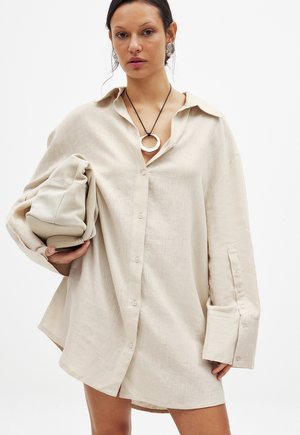 Lichtbeige linnen shirt met een knoopsluiting aan de voorkant, lange mouwen en een omslagkraag. Model houdt een bijpassend zacht stoffen item vast.