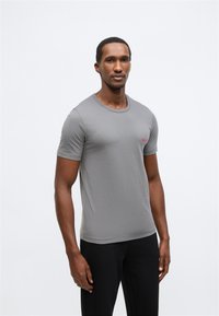 Šedé tričko s krátkým rukávem a kulatým výstřihem, s malým červeným logem na levé straně hrudi, vyrobené z hladké tkaniny, doplněné o texturované černé kalhoty.