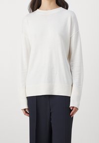 Pullover lungo a maniche, crema chiaro, con scollo e polsini a coste. Presenta una vestibilità rilassata e una texture morbida, abbinato a pantaloni scuri.