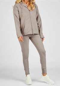 Jasnobrązowy dwuczęściowy dres z bluzą na zamek z kieszenią z przodu oraz dopasowanymi zwężanymi spodniami. Noszony z białymi trampkami.