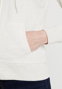 Sudadera de algodón gris claro con un bolsillo frontal tipo canguro y puños acanalados. La tela tiene una textura suave con un sutil patrón jaspeado.
