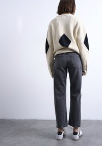 Άτομο που στέκεται με την πλάτη  γυρισμένη, φοράει μπεζ πουλόβερ με μαύρα διαμαντένια σχέδια, σκούρα γκρι τζιν και λευκά sneakers σε τσιμεντένιο δάπεδο.