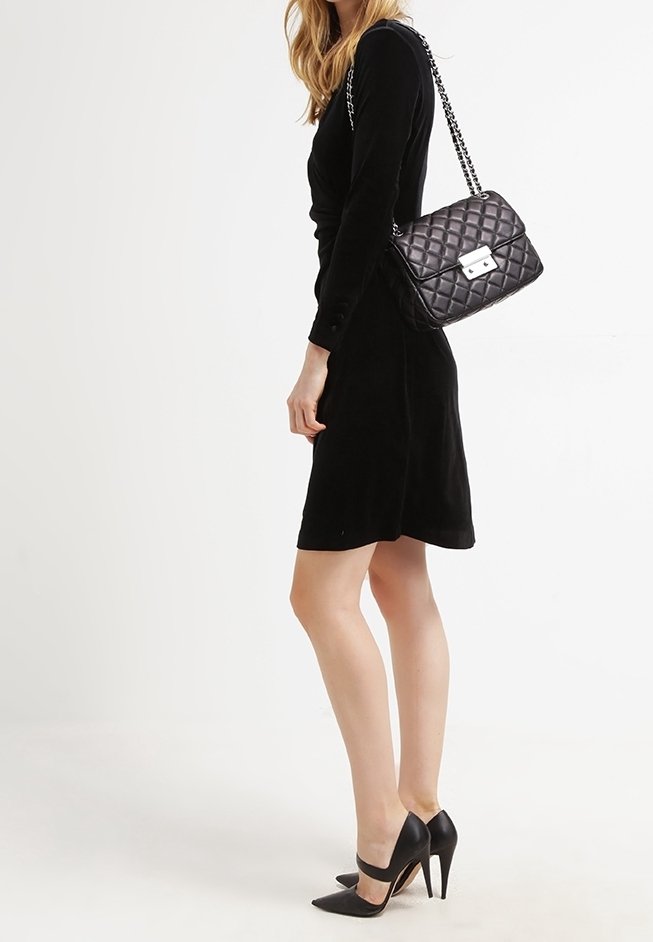 Femme portant une robe noire à manches longues, des escarpins noirs et un sac à main matelassé noir avec une bandoulière chaîne sur l'épaule.