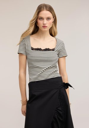 Jeune femme aux longs cheveux blonds portant un haut rayé noir et blanc avec un détail en dentelle et une jupe portefeuille noire avec un nœud sur le côté.