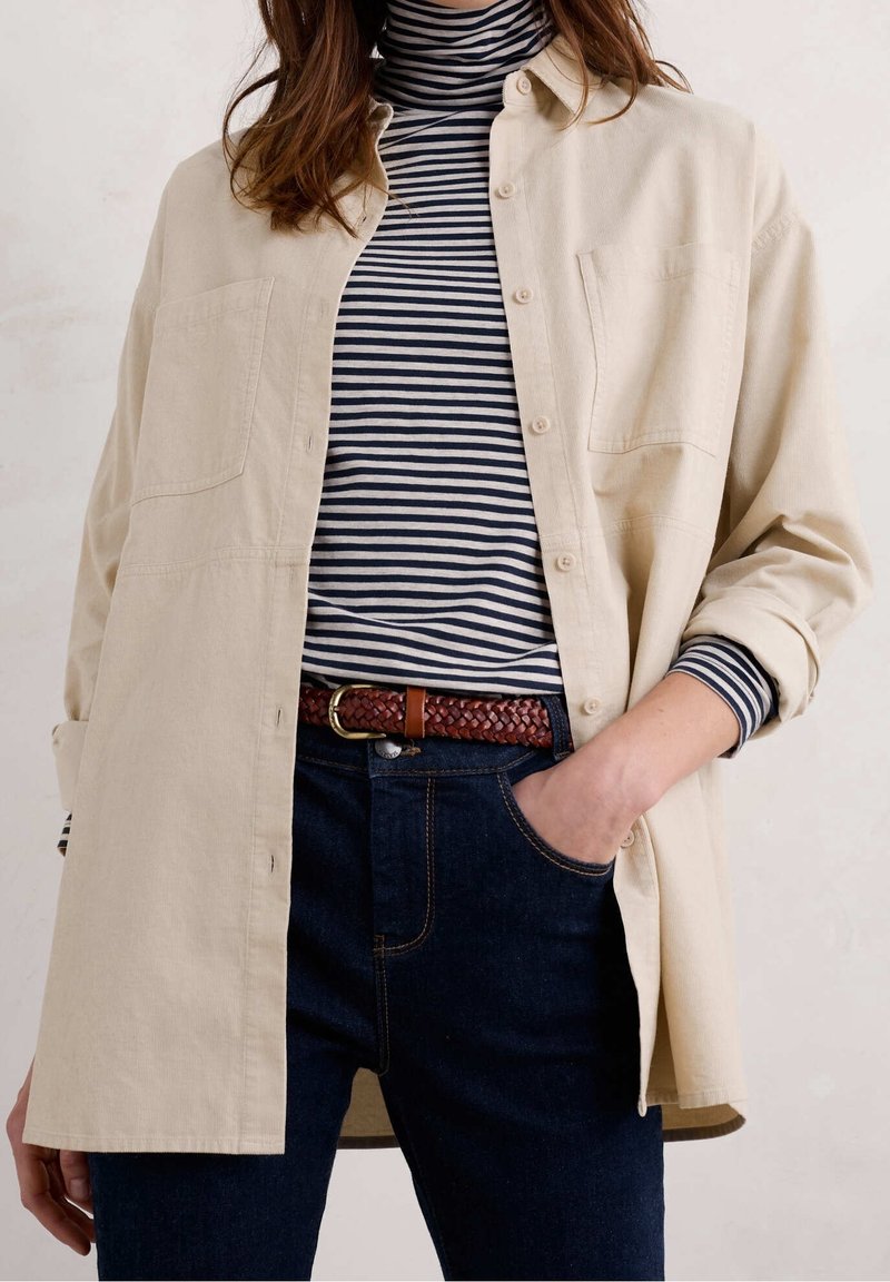 Beige button-up shirt over a navy and white striped turtleneck, paired with dark jeans and a brown braided belt, hands in pockets.