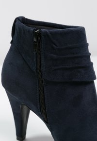 Granatowy zamszowy botek sięgający kostki z bocznym zamkiem błyskawicznym i marszczeniem przy kołnierzu. Posiada szpilkowy obcas i gładką fakturę.