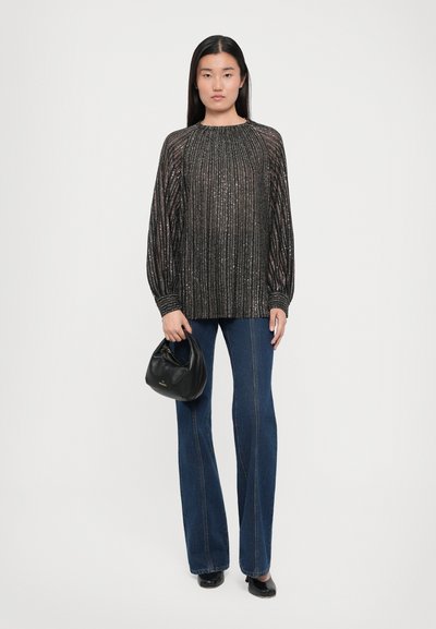 Top lungo a maniche lunghe plissettato nero con filo metallico, abbinato a jeans a zampa d'elefante blu. La modella tiene una piccola borsa nera.