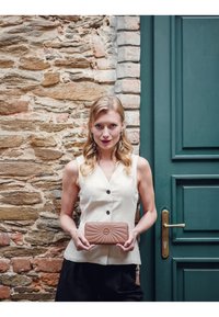 Beige quilted wallet with a patterned design and circular emblem, held in two hands against a stone wall backdrop.