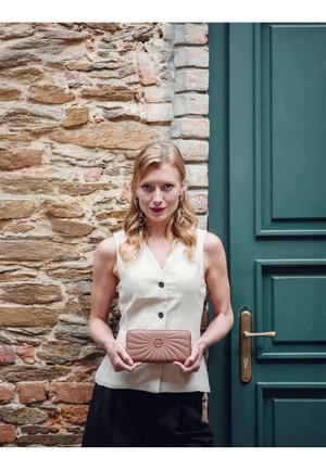 Beige quilted wallet with a patterned design and circular emblem, held in two hands against a stone wall backdrop.