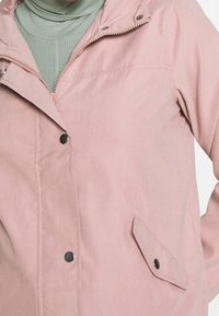 Ljusrosa jacka med dragkedja och tryckknappar. Den har en mjuk textur, en bröstficka fram och en huva.